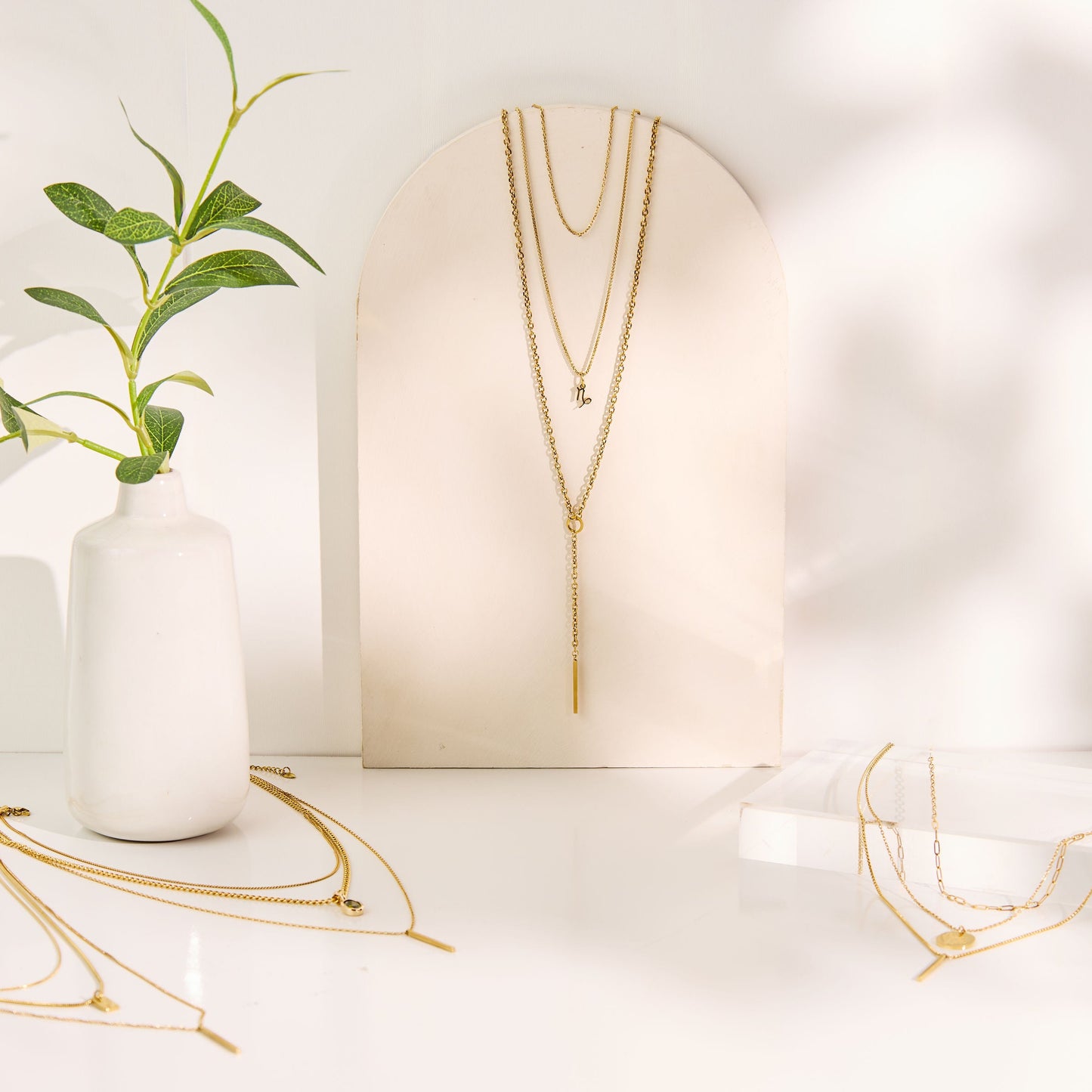 Gold necklaces displayed on a white surface with a vase and plant in the background